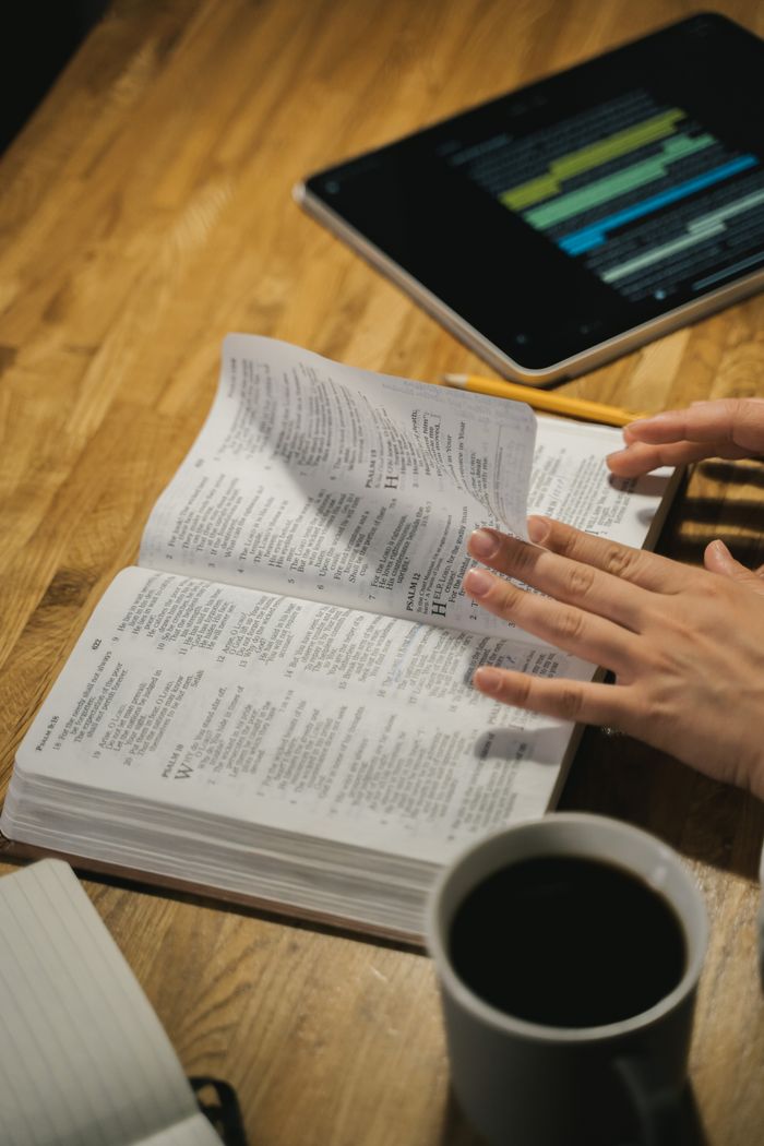 Offerings Close-up of hands turning Bible pages with a coffee cup and tablet on the wooden table.